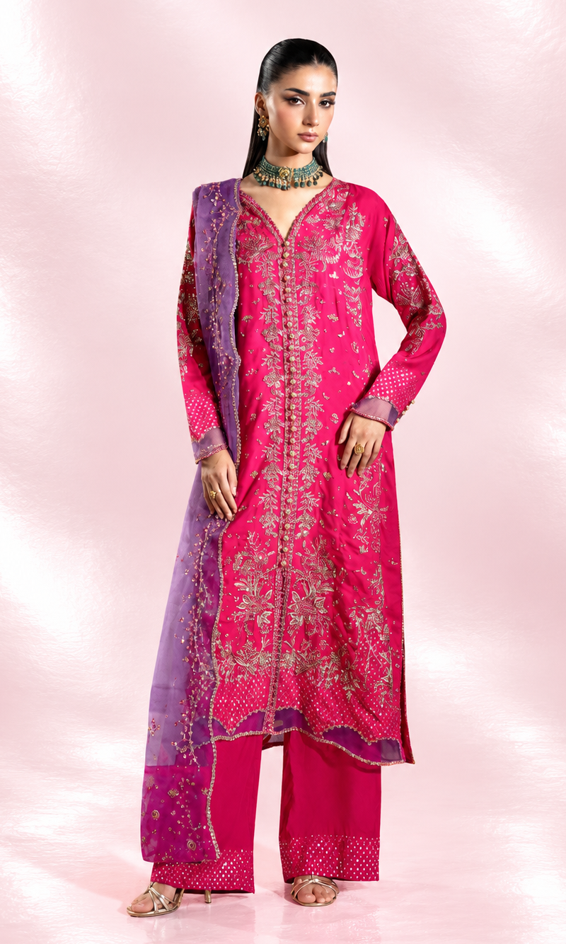 A woman modeling a bright pink traditional outfit with gold embroidery, matching pink pants, and a purple dupatta with gold detailing, set against a pink studio backdrop.