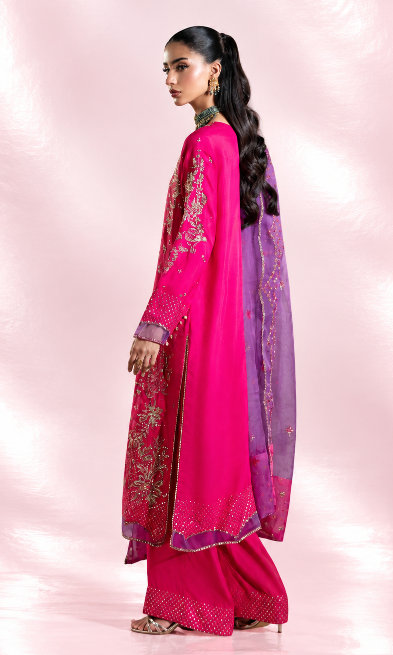 A woman modeling a traditional pink and purple embroidered outfit with a matching sheer purple dupatta, standing against a soft pink studio backdrop.