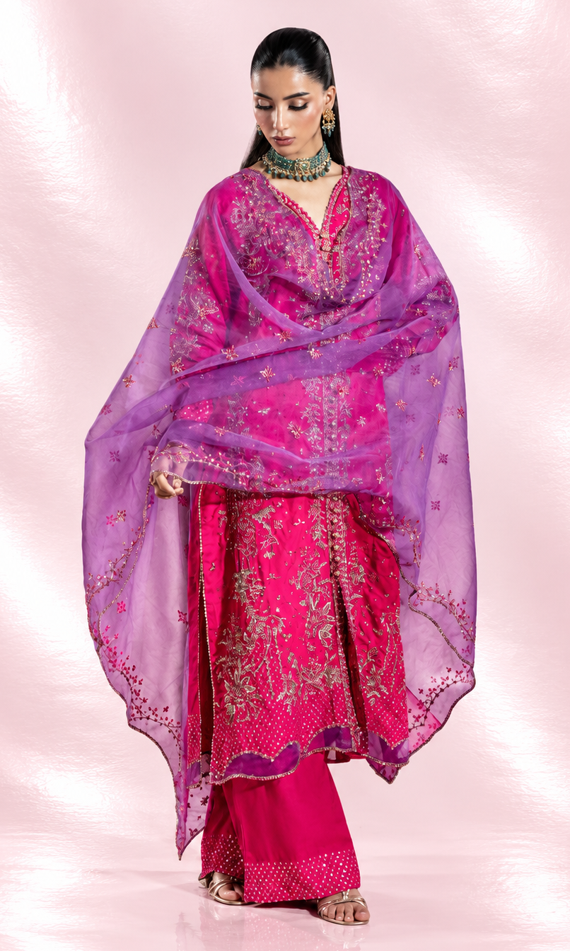 A woman models a traditional Indian outfit featuring a bright pink kurta with gold embroidery, matching pink pants, and a sheer purple dupatta with gold floral patterns, set against a pink textured studio backdrop.