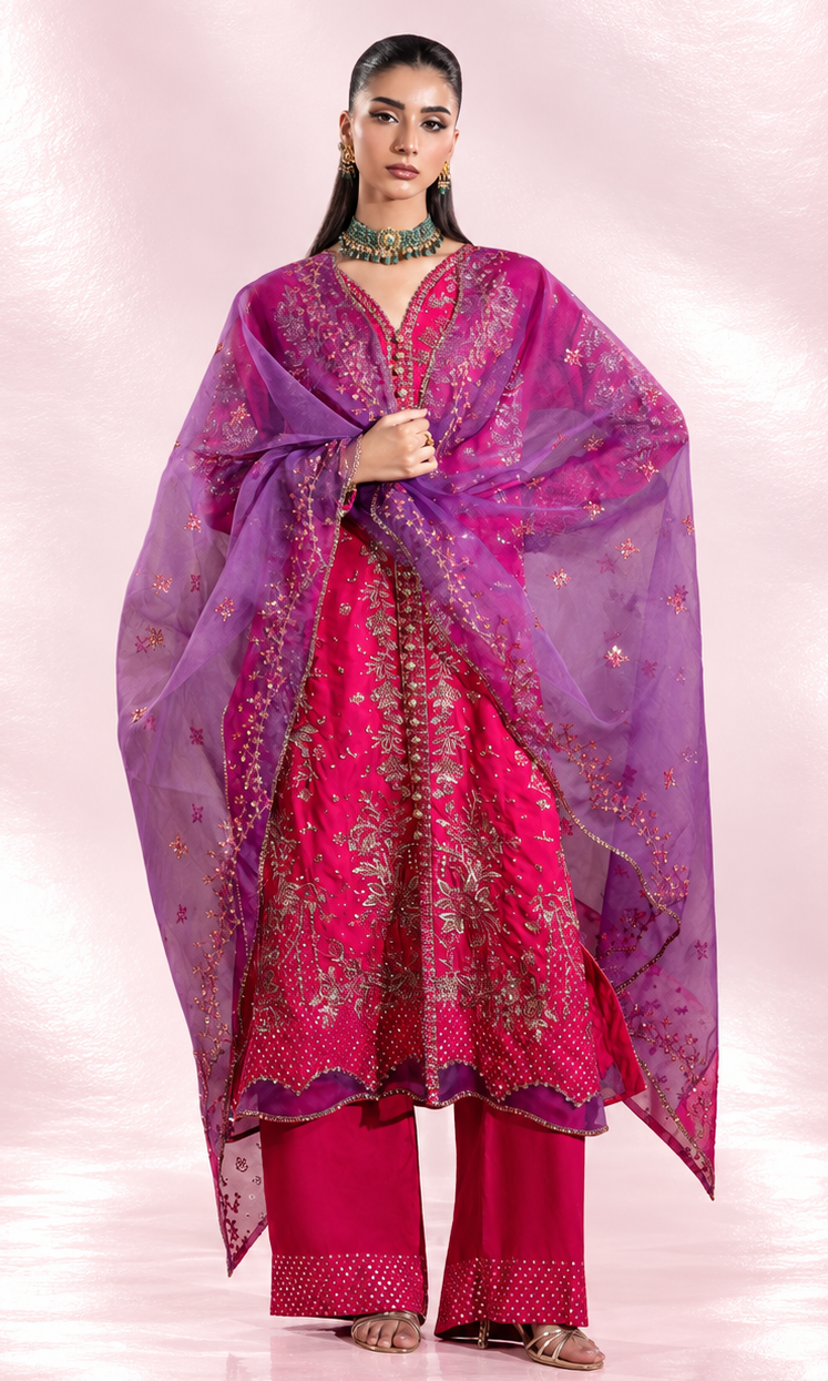 A woman modeling a traditional Indian outfit consisting of a bright pink embroidered kurta, matching pink pants, and a sheer purple dupatta with gold embroidery, set against a pink studio backdrop.