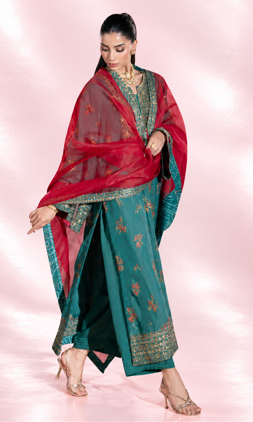 A woman models a traditional Indian outfit consisting of a teal green salwar kameez with gold embroidery, a red dupatta with gold motifs, and a black inner skirt, set against a pink studio backdrop.