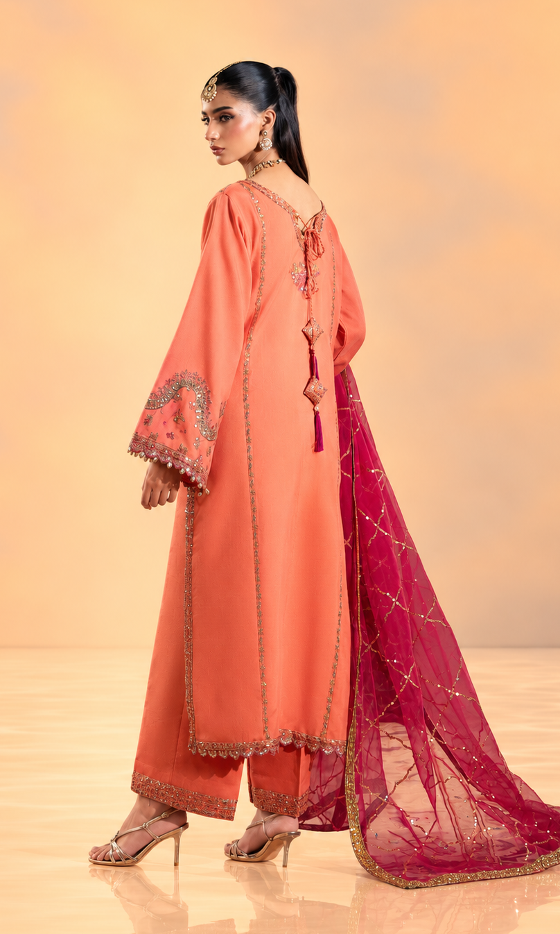 A woman modeling a peach-orange traditional outfit with matching pants and a deep magenta dupatta featuring gold embroidery, set against a warm beige studio background.