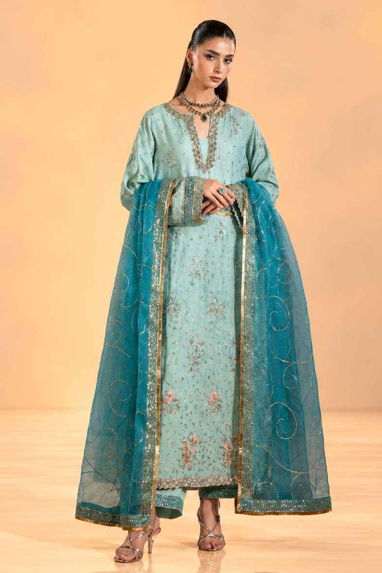 A woman modeling a traditional long teal anarkali-style dress with floral embroidery and a matching sheer dupatta, photographed against a warm beige studio backdrop.