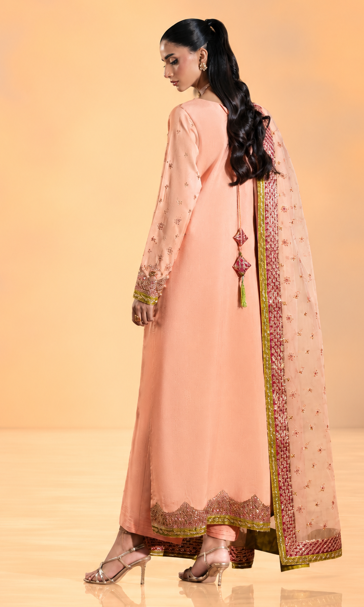 A woman modeling a peach-pink traditional Anarkali kurta with a matching printed dupatta, shown from a side-back angle against a warm beige studio backdrop.