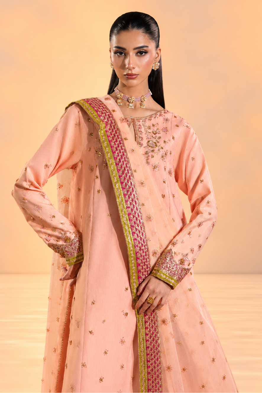 A woman modeling a peach-pink traditional South Asian outfit with gold embroidery and a patterned green and pink border dupatta, set against a warm beige studio background.