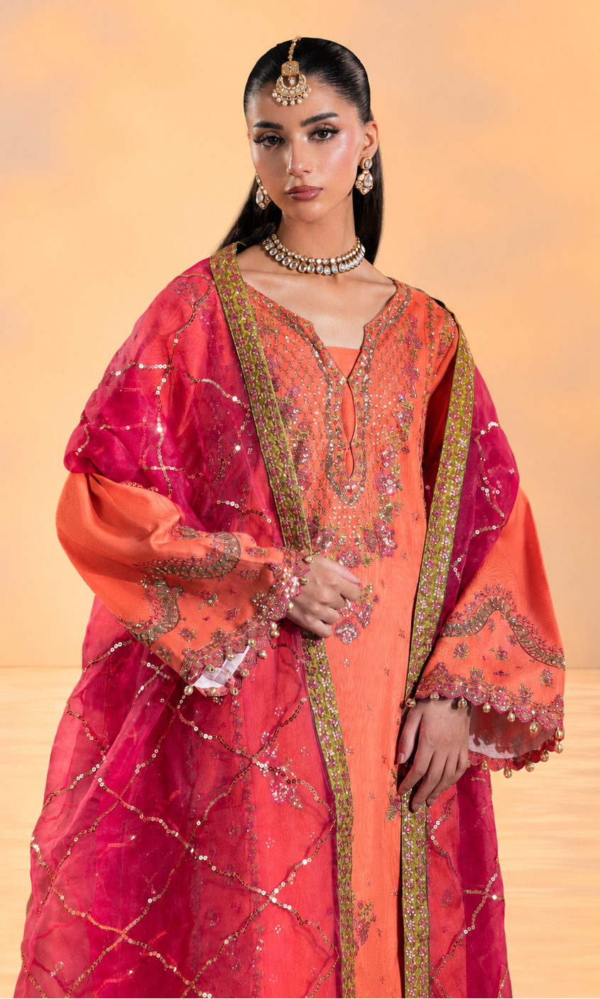 A woman modeling a traditional Indian outfit consisting of a bright orange kurta with gold embroidery, a pink dupatta with gold geometric patterns, and a matching pink outer layer, set against a warm beige background.