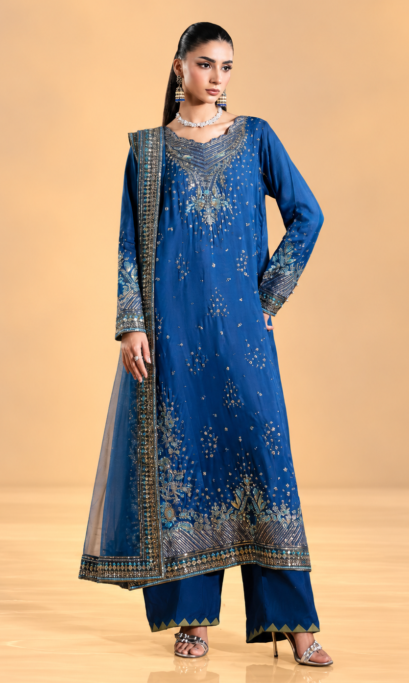 A woman modeling a royal blue traditional South Asian outfit with intricate gold and silver embroidery, matching wide-leg pants, and a sheer dupatta, set against a warm beige studio background.