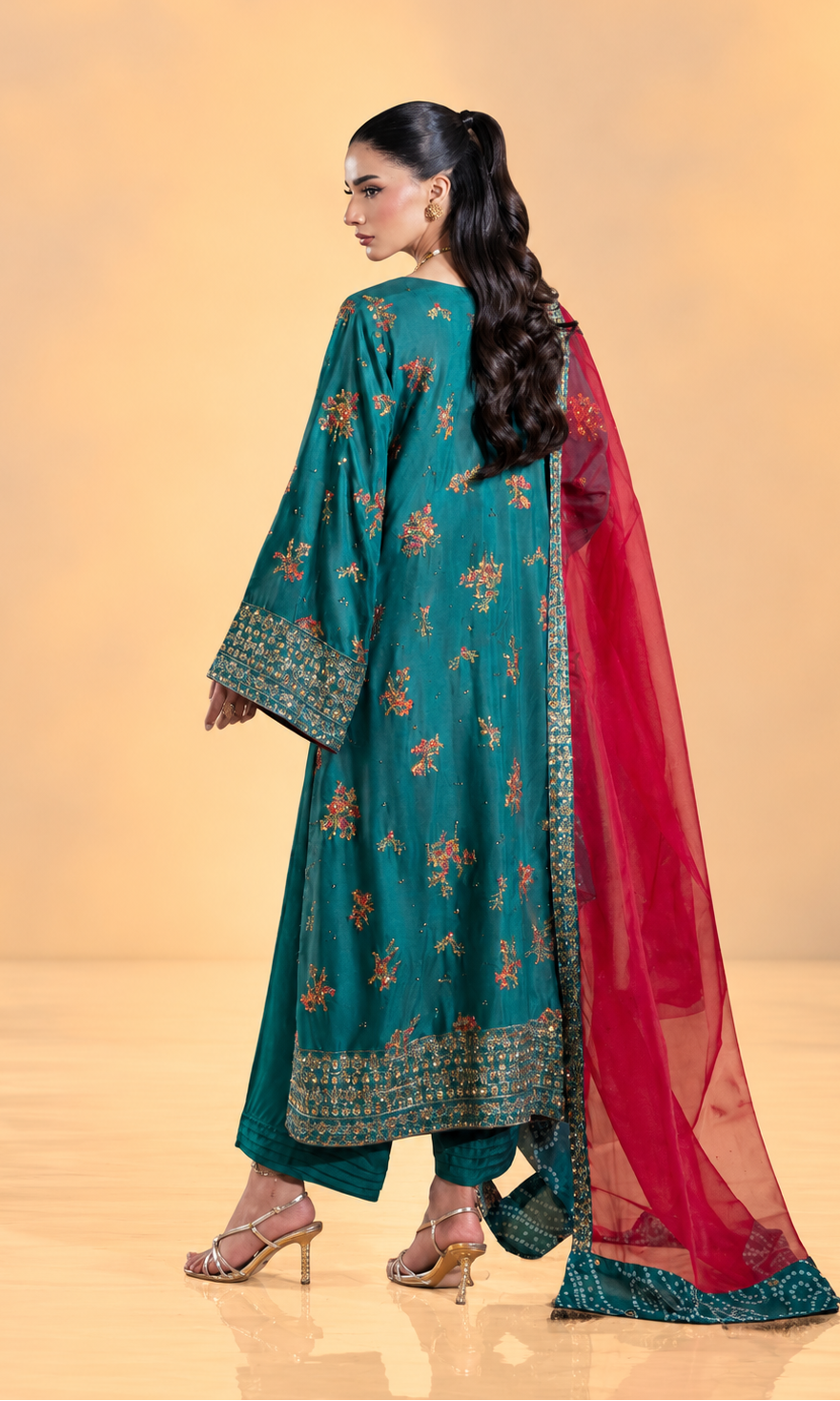 A woman modeling a traditional teal green salwar kameez suit with floral embroidery, a matching green dupatta, and a bright red draped overlay, shown from the back against a warm beige studio background.