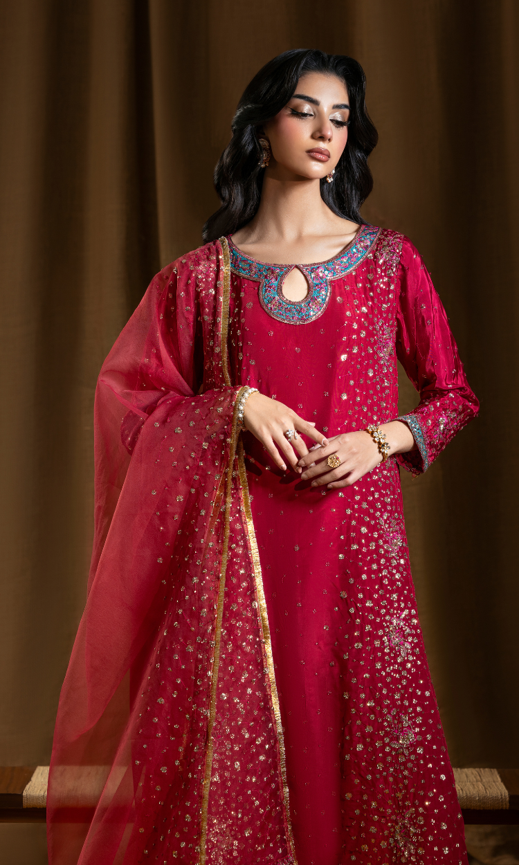 A woman modeling a traditional Indian salwar kameez outfit in a rich magenta pink fabric adorned with gold and silver sequins and embroidery. The outfit includes a long kurta with a decorative embroidered neckline, matching flared sleeves, and a sheer dupatta draped over her shoulder, set against a dark brown draped background.